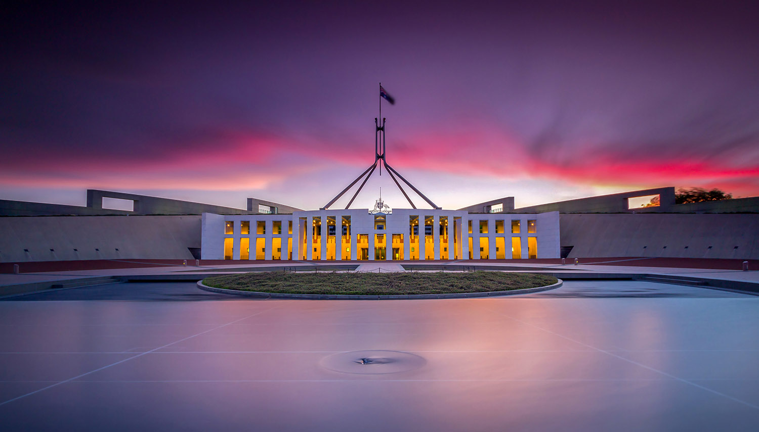 A short photo all the Australian Parliament House at dusk with beautiful pinks and purples from the sky reflected in the lake and the windows lit up