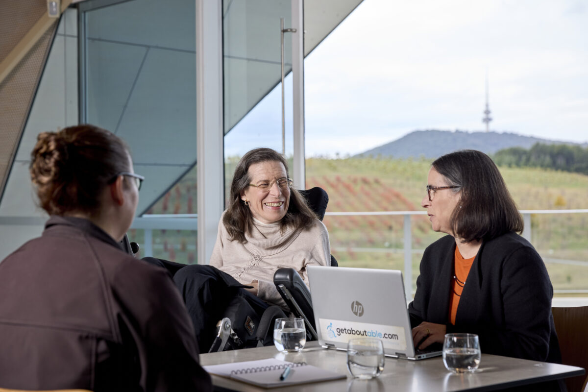 3 people at a table with a laptop. one is facing away from camera and 1 is a wheelchair user,